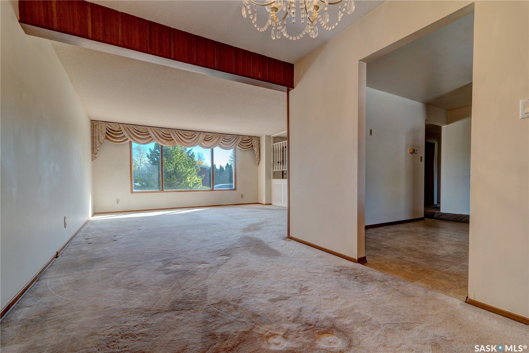 Arched window valance, large multi-pane window, open concept room division, chandelier fixture, and wood-paneled ceiling beam