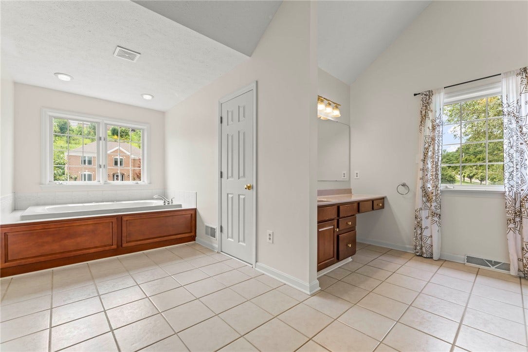 The luxurious primary bathroom offers a soaring cathedral ceiling, a jetted soaking tub, separate shower, dual vanities positioned on opposite walls for added convenience, and an additional storage area.